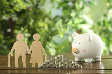 Photo of Pension savings. Figure of senior couple, piggy bank and stacked coins on wooden table outdoors