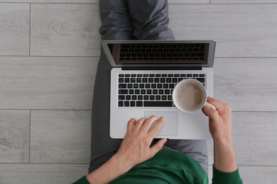 Photo of Young man with cup of coffee using laptop while sitting on floor, top view