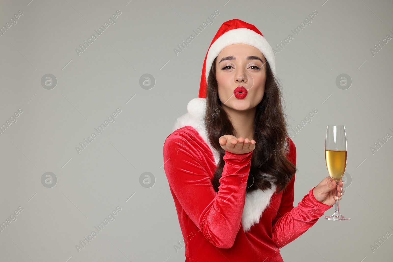 Photo of Christmas celebration. Beautiful young woman in red dress and Santa hat with glass of champagne blowing kiss on grey background, space for text