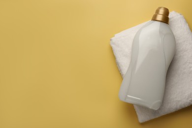 Photo of Bottle of fabric softener and terry towel on pale yellow background, top view. Space for text