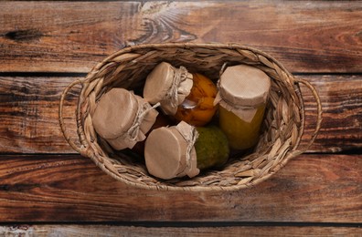 Photo of Wicker basket with many jars of different preserved products on wooden table, top view