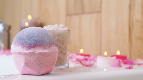 Photo of Colorful bath bomb, sea salt, flower petals and burning candles on white tub in bathroom, closeup. Space for text