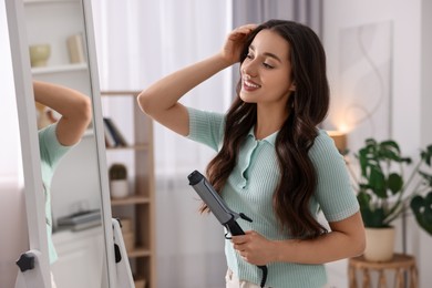 Photo of Smiling woman with beautiful hairstyle holding curling hair iron near mirror at home