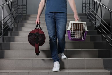 Photo of Travel with pet. Man holding carrier with cute cat and bag on stairs indoors, closeup