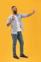 Smiling young man taking selfie with smartphone and showing peace sign on yellow background