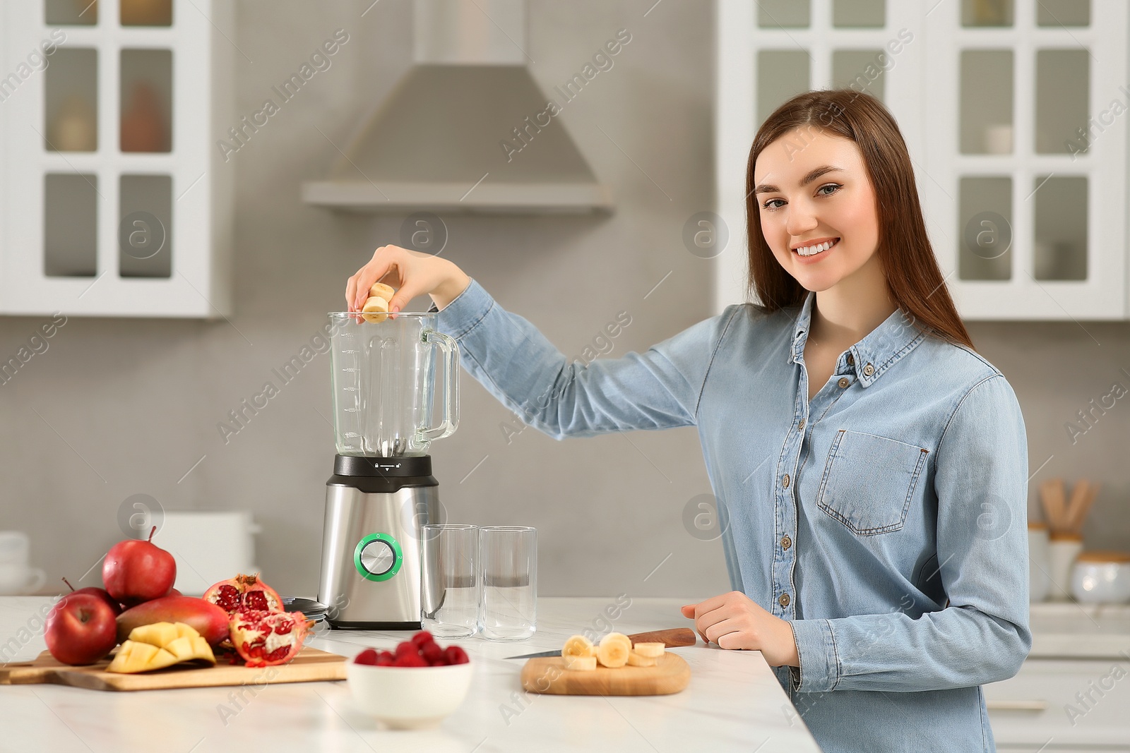 Photo of Beautiful young woman adding banana into blender for tasty smoothie in kitchen