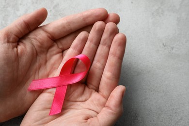 Woman holding pink ribbon at grey table, closeup with space for text. Breast cancer awareness concept