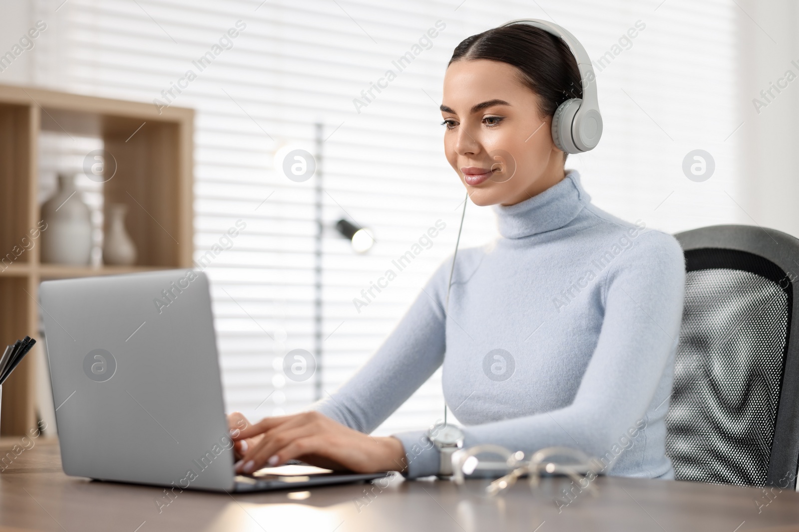 Photo of Young woman in headphones watching webinar at table in office