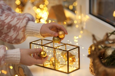 Photo of Christmas atmosphere. Woman putting beautiful baubles into decorative container on window sill indoors, closeup