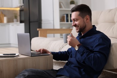 Happy man with cup of drink working on laptop at wooden coffee table in room