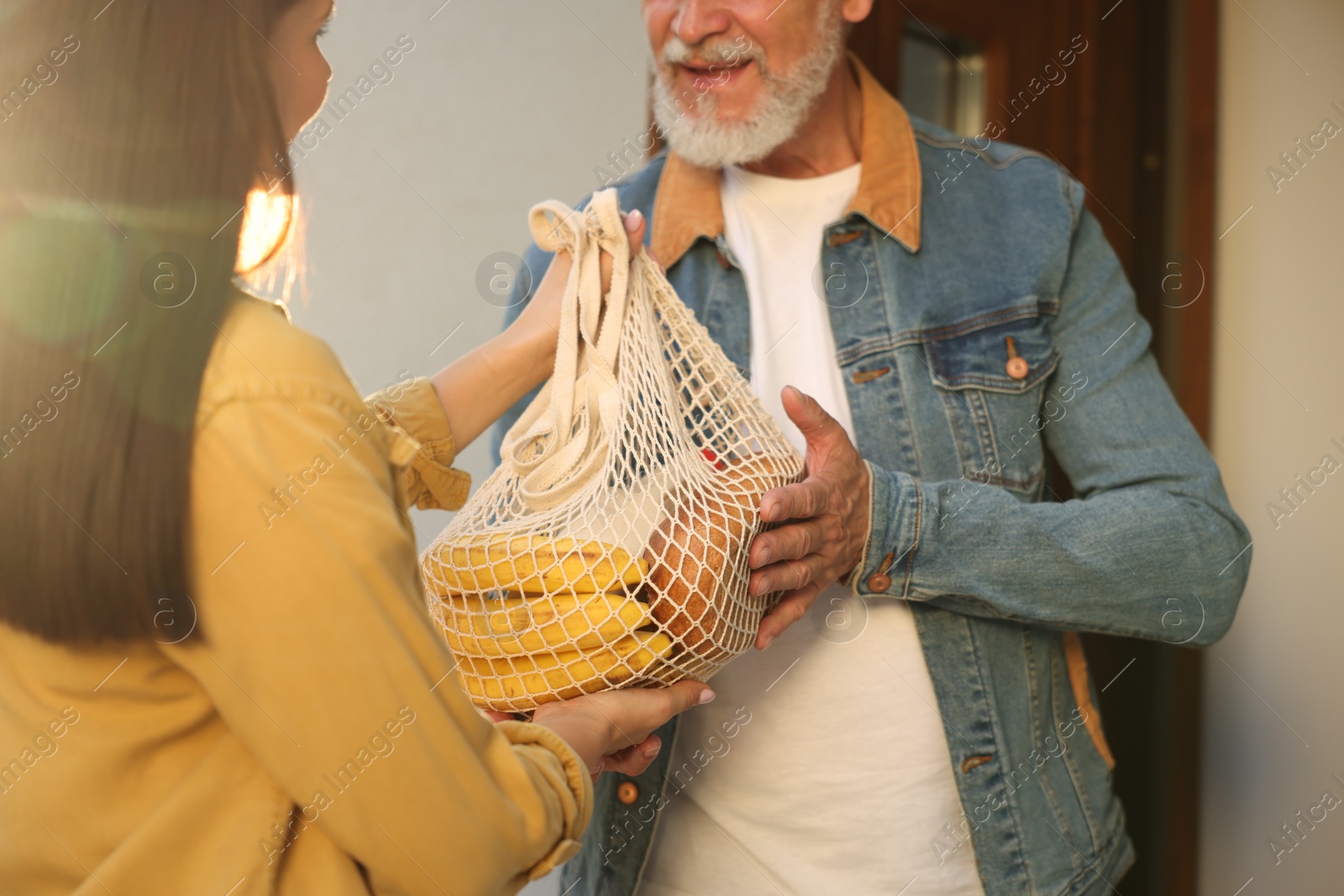 Photo of Helping neighbours. Young woman with net bag of products visiting senior man outdoors on sunny day, closeup