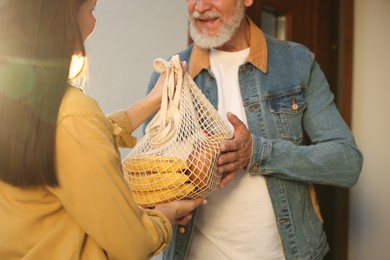 Photo of Helping neighbours. Young woman with net bag of products visiting senior man outdoors on sunny day, closeup