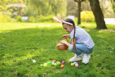 Photo of Easter celebration. Cute little girl in bunny ears hunting eggs outdoors, space for text