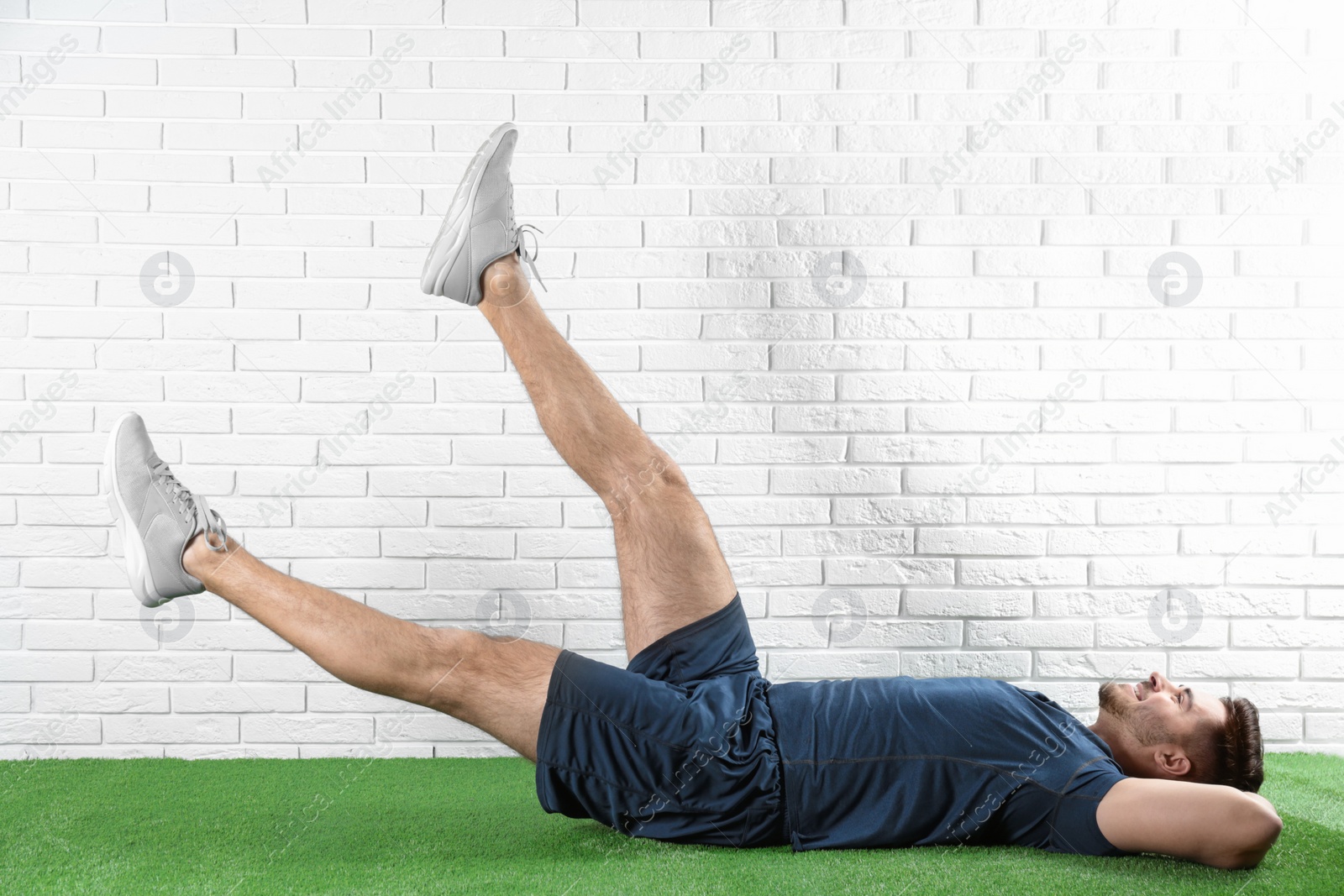 Photo of Handsome young man in sportswear doing scissors exercise on artificial green lawn near brick wall