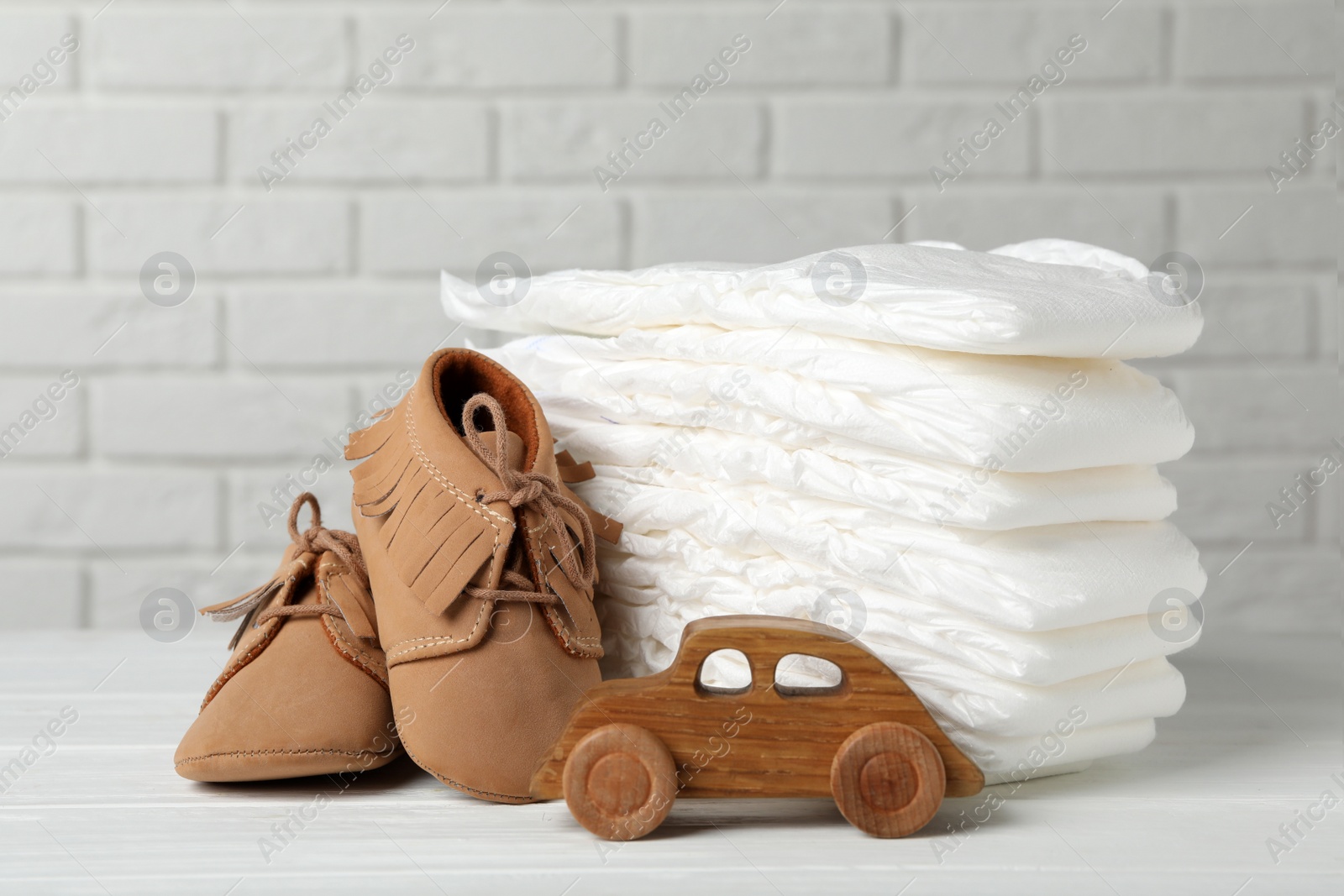Photo of Baby diapers, toy car and child's shoes on wooden table against white brick wall