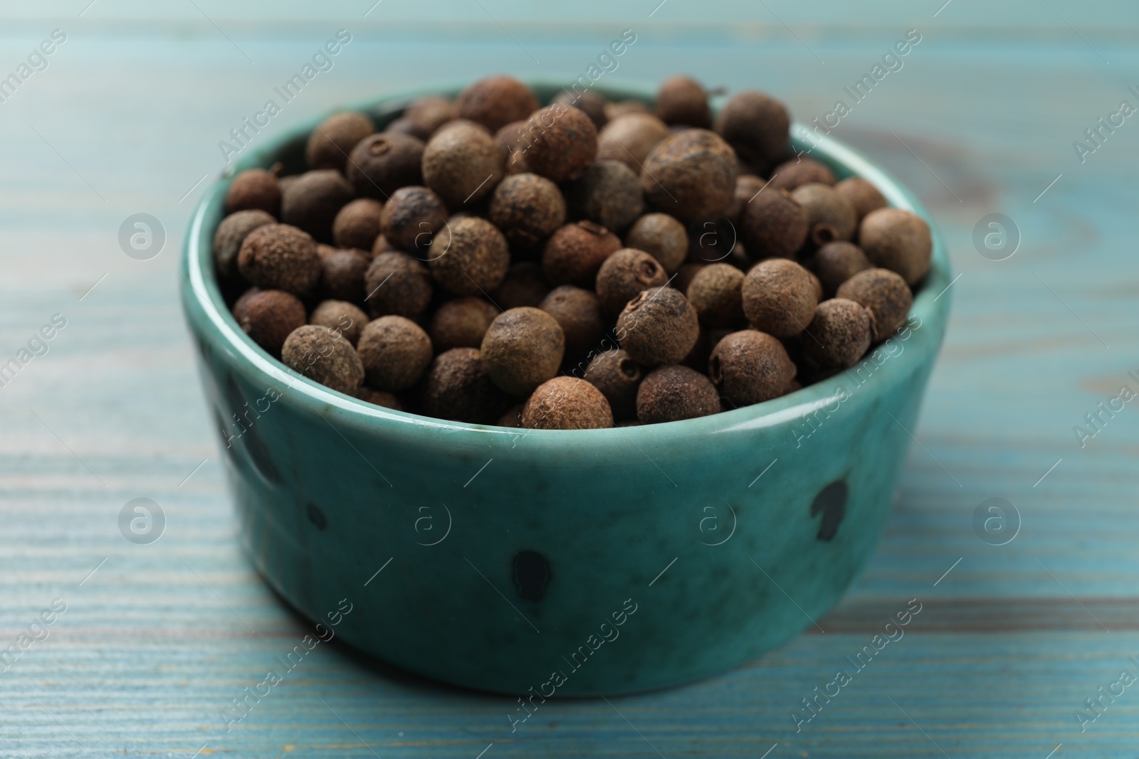 Photo of Aromatic allspice pepper grains in bowl on light blue wooden table, closeup