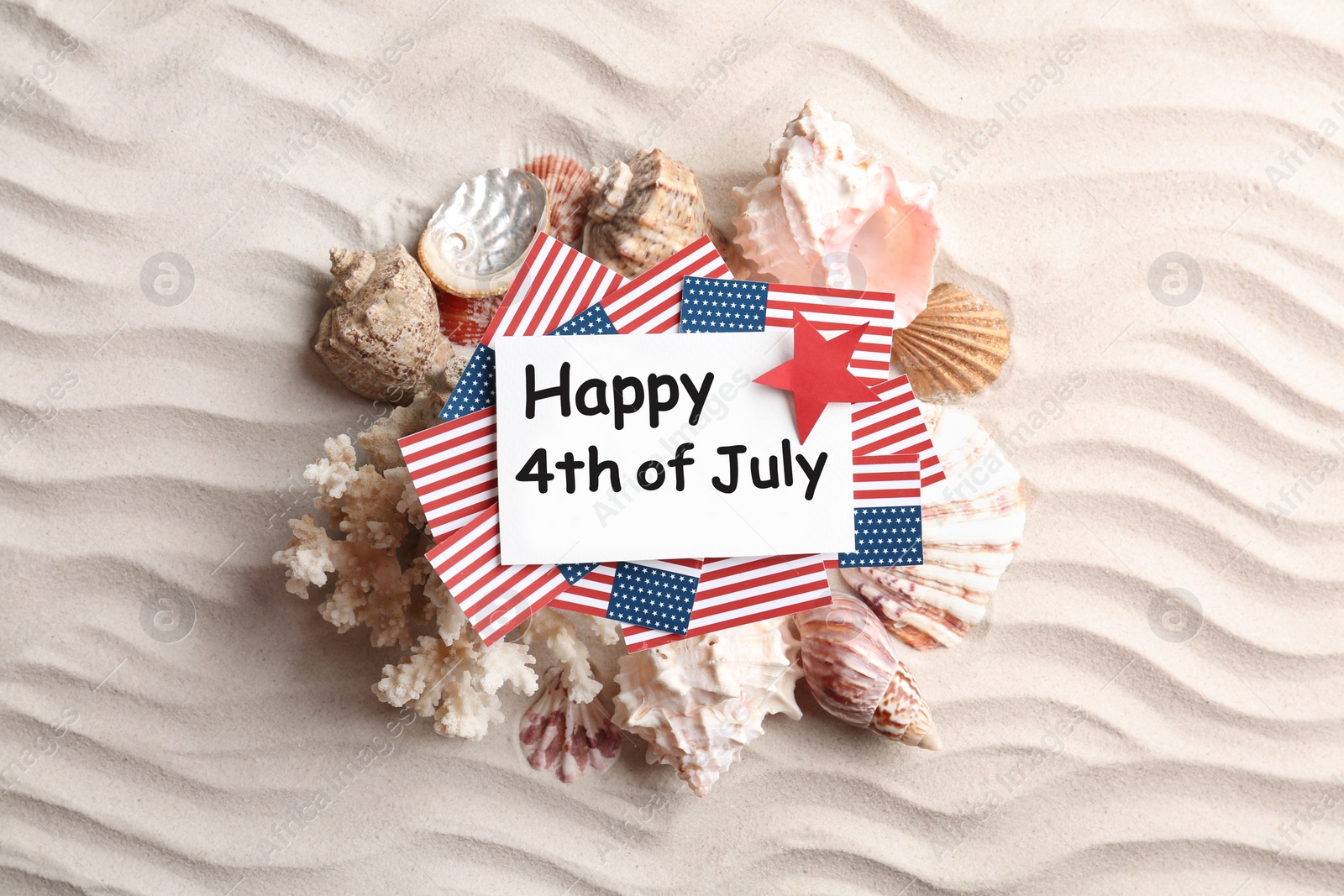 Photo of Flat lay composition with greeting card, USA flags and seashells on sand. Happy Independence Day