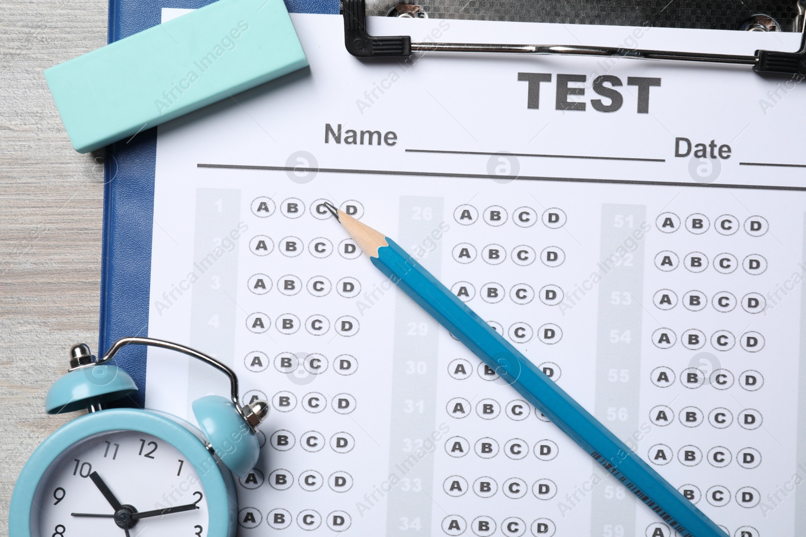 Photo of Clipboard with answer sheet, pencil and alarm clock on light wooden table, flat lay. Student passing exam