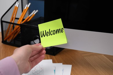 Image of Woman taking sticky note with word Welcome from computer monitor at office desk, closeup