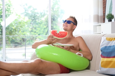 Shirtless man with inflatable ring eating watermelon on sofa at home