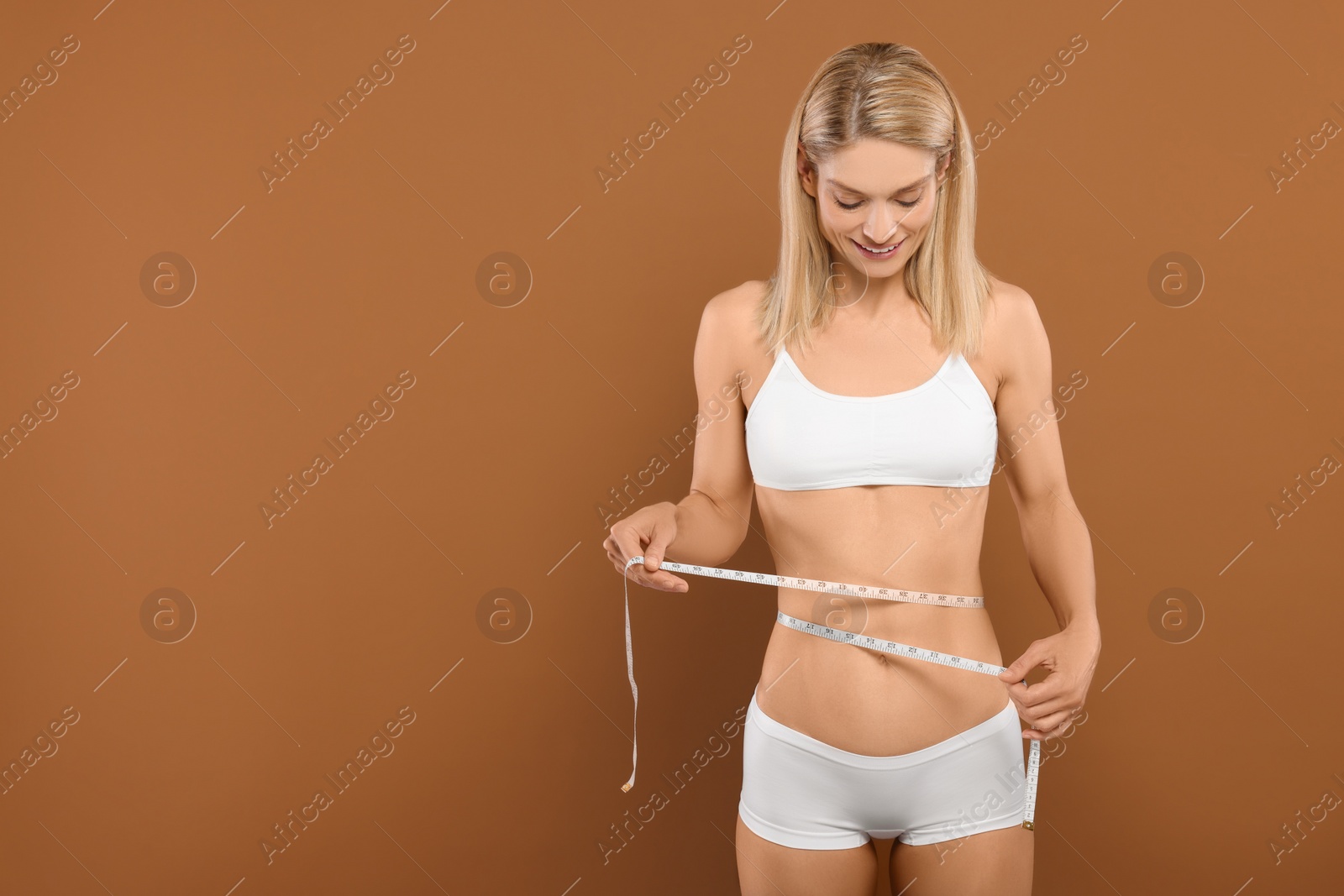 Photo of Slim woman measuring waist with tape on brown background, space for text. Weight loss