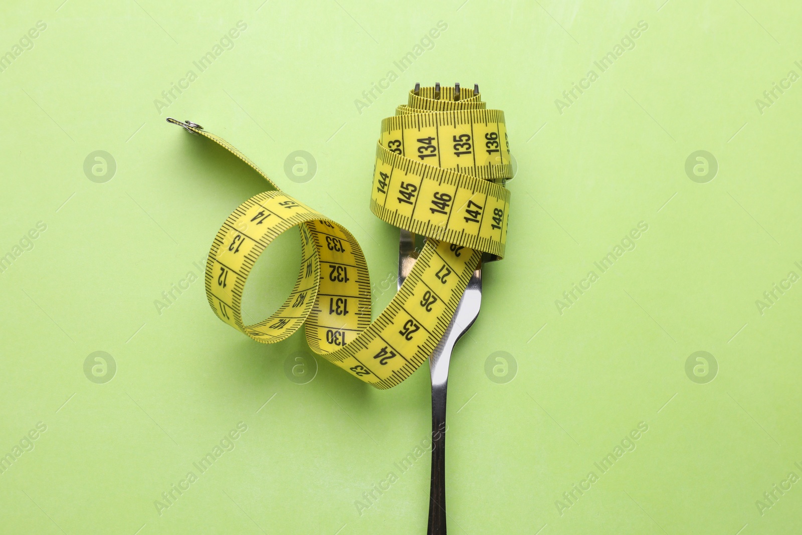 Photo of Fork with measuring tape on light green background, top view. Diet concept