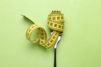 Photo of Fork with measuring tape on light green background, top view. Diet concept