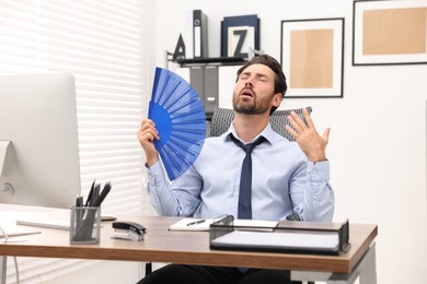 Bearded businessman waving blue hand fan to cool himself at table in office