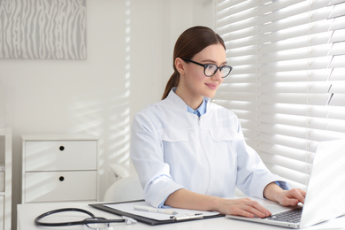 Photo of Young female doctor working with laptop at table in office