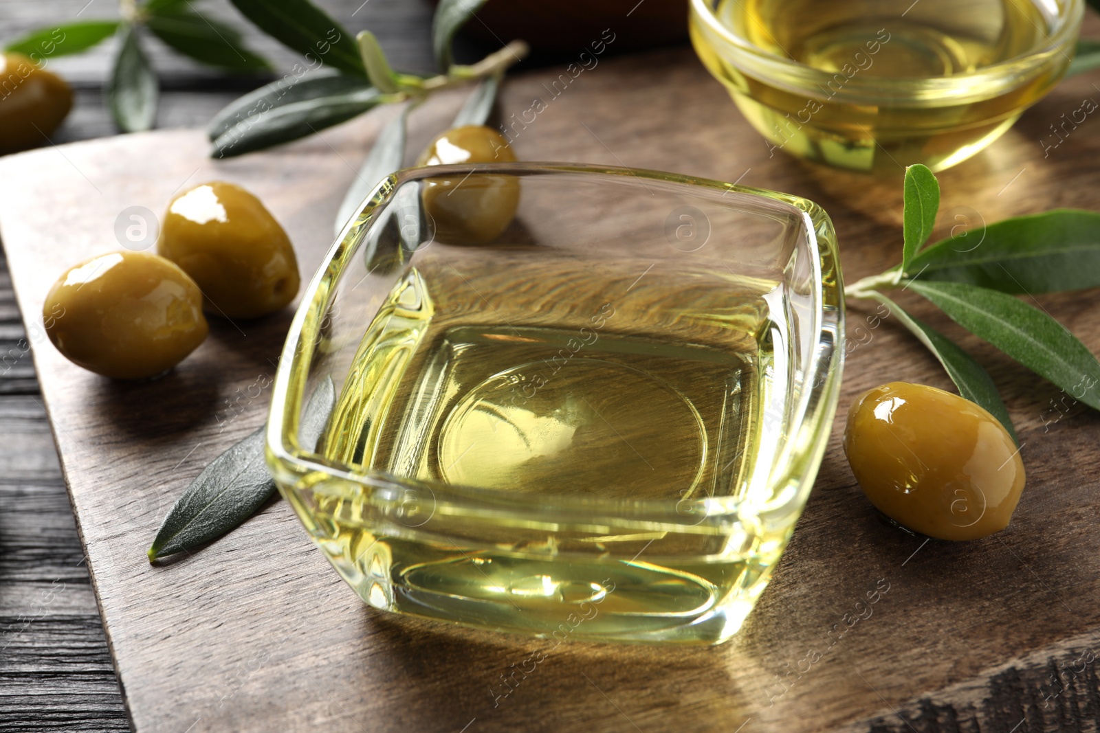 Photo of Glass bowls of oil, ripe olives and green leaves on black wooden table, closeup