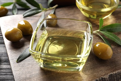 Photo of Glass bowls of oil, ripe olives and green leaves on black wooden table, closeup