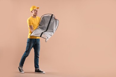 Photo of Dry-cleaning delivery. Happy courier holding garment cover with clothes on beige background, space for text