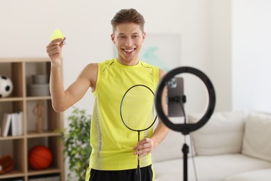 Photo of Smiling sports blogger holding badminton racket and shuttlecock while streaming online fitness lesson with smartphone at home