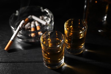 Photo of Alcohol addiction. Whiskey in glasses, smoldering cigarette and ashtray on black wooden table, closeup