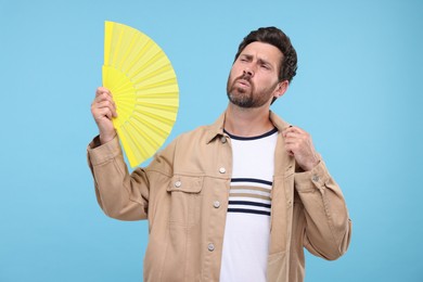 Photo of Unhappy man with hand fan suffering from heat on light blue background
