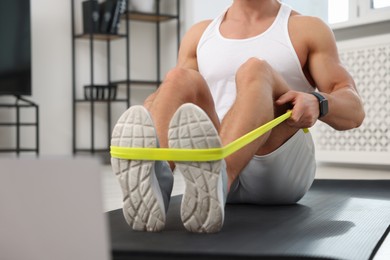 Photo of Muscular man doing exercise with elastic resistance band near laptop on mat at home, closeup