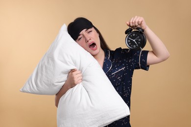 Photo of Tired young woman with sleep mask and pillow yawning on beige background. Insomnia problem