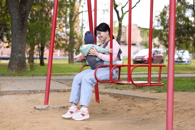 Mother holding her child in sling (baby carrier) on swing in park
