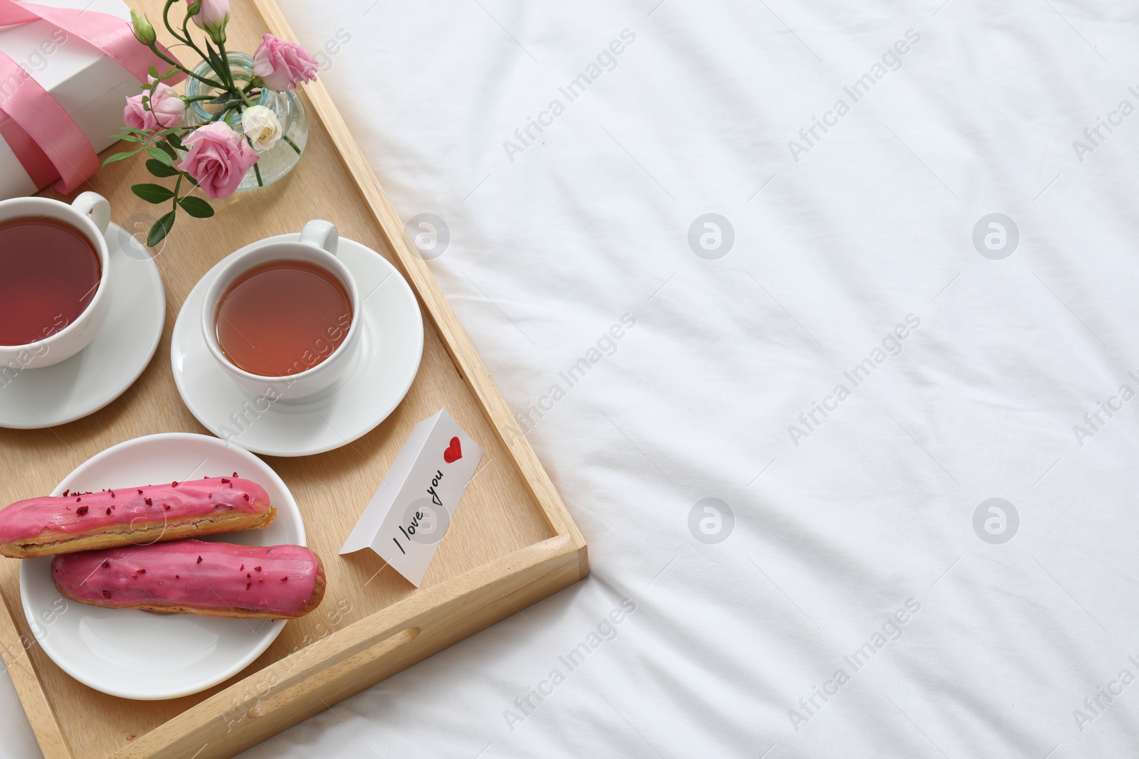 Photo of Tasty breakfast served in bed. Delicious eclairs, tea, gift box, flowers and card with phrase I Love You on tray, above view. Space for text