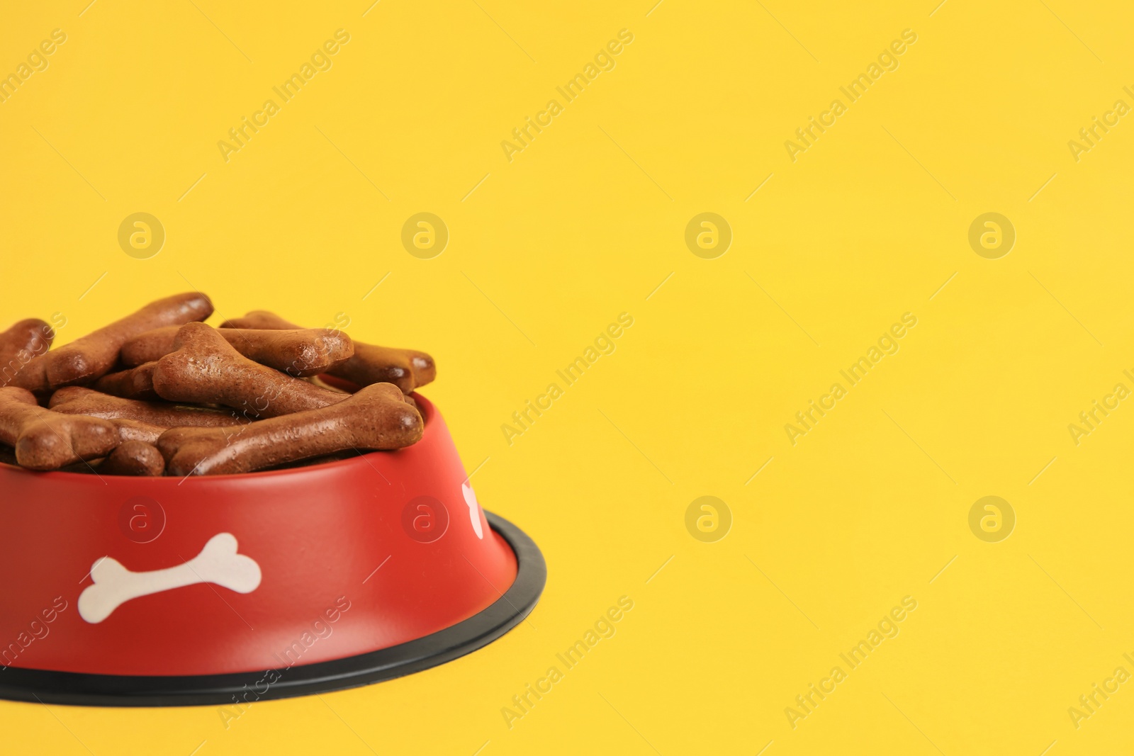 Photo of Red bowl with bone shaped dog cookies on yellow background, closeup. Space for text