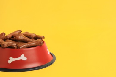Photo of Red bowl with bone shaped dog cookies on yellow background, closeup. Space for text