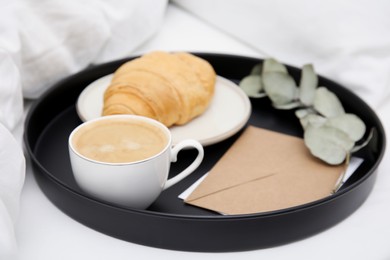 Photo of Tray with tasty croissant, cup of coffee and envelope on white bed