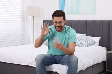 Photo of Happy man greeting someone during video chat via smartphone in bedroom