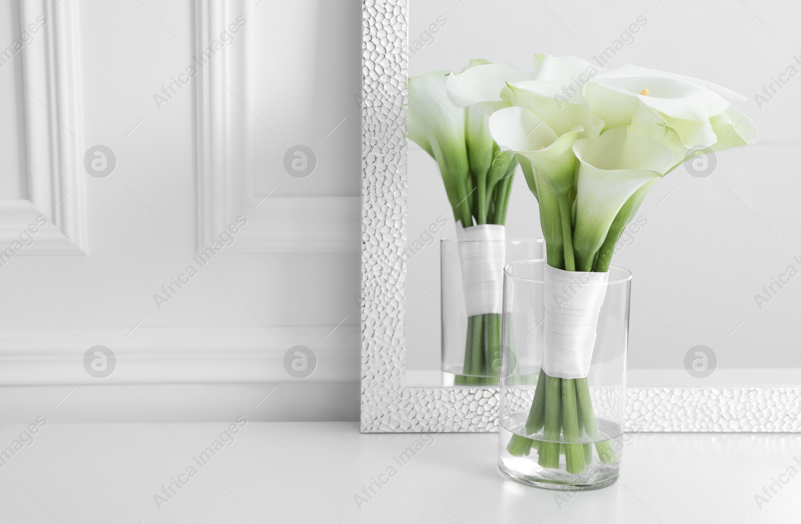 Photo of Beautiful calla lily flowers tied with ribbon in glass vase on white table, space for text