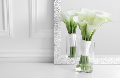 Photo of Beautiful calla lily flowers tied with ribbon in glass vase on white table, space for text
