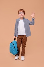 Happy schoolboy with backpack waving hello on beige background