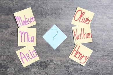 Paper notes with different baby names and question mark on grey table, flat lay