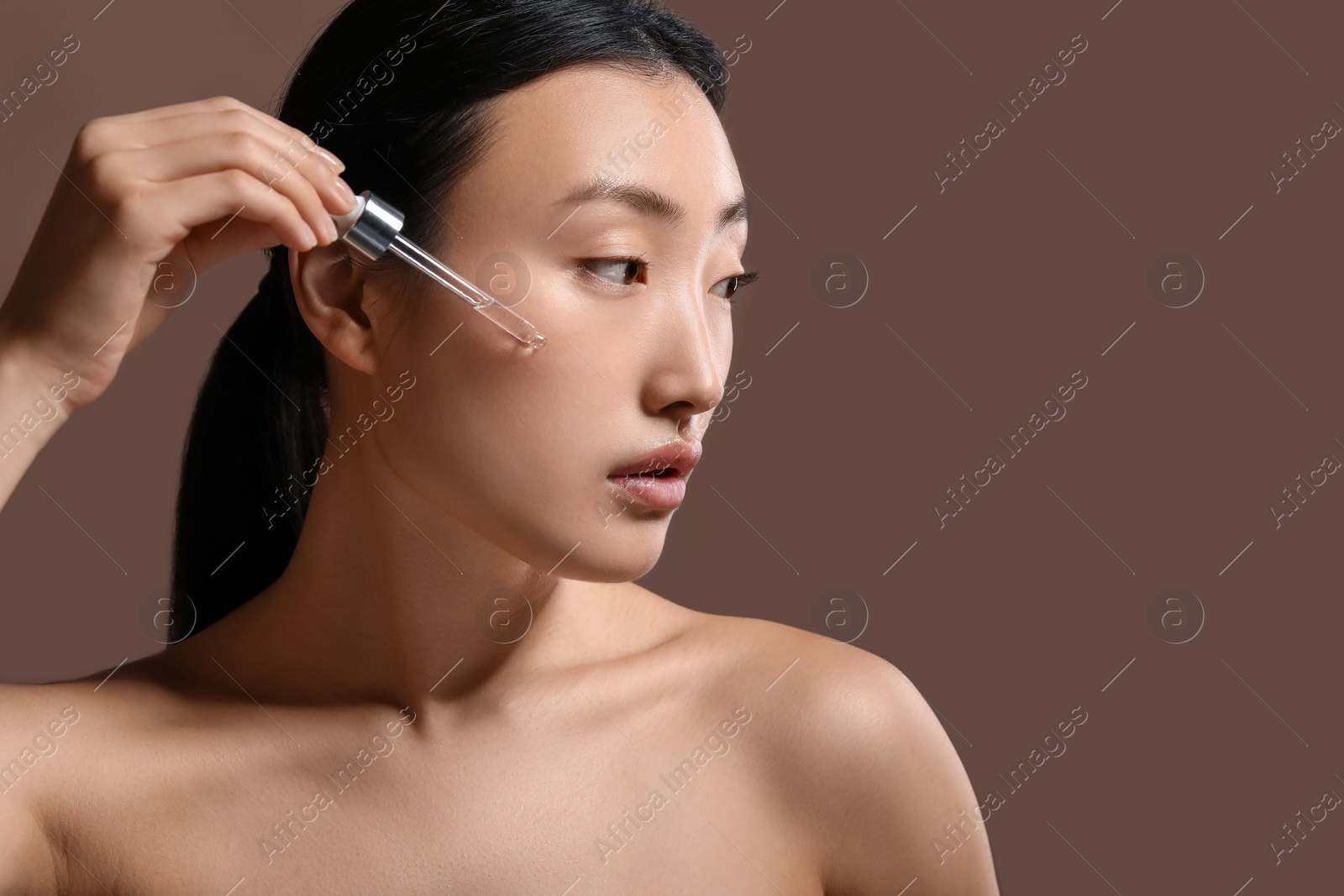 Photo of Beautiful young woman applying cosmetic serum onto her face on brown background, space for text