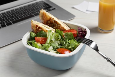 Photo of Container of tasty food, laptop, fork and glass of juice on white wooden table. Business lunch