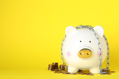 Photo of Piggy bank with steel chain and coins on yellow background, space for text. Money safety concept